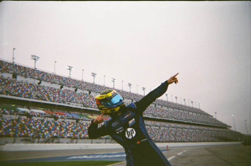 Un piloto de carreras con traje azul y casco amarillo posa para celebrar en la pista frente a las gradas vacías del estadio.