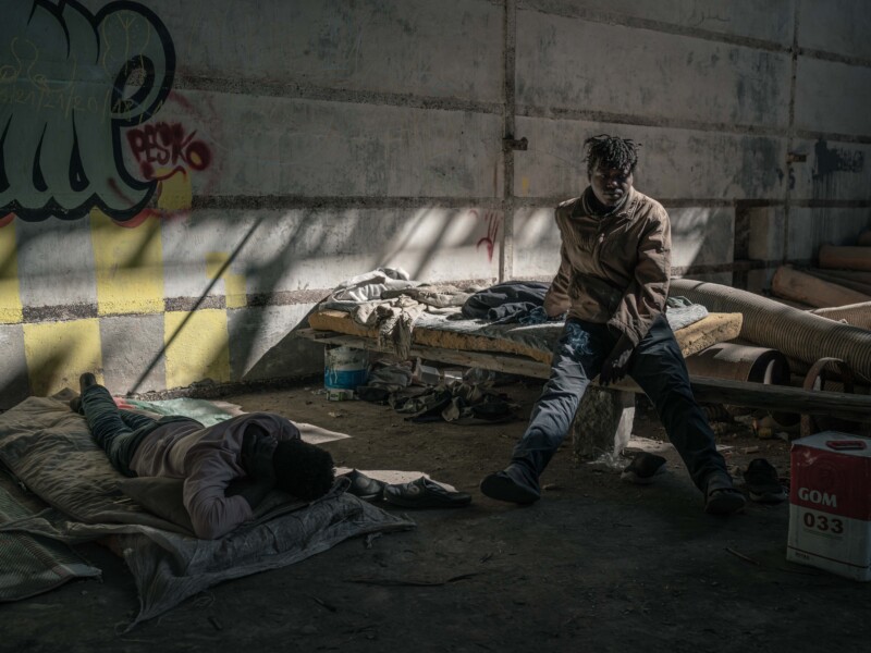 Two people in worn clothes rest inside an abandoned, dimly lit building. One person lies on a blanket on the floor, while the other sits on a broken bed. Sunlight streams in, highlighting the graffiti-covered wall behind them.