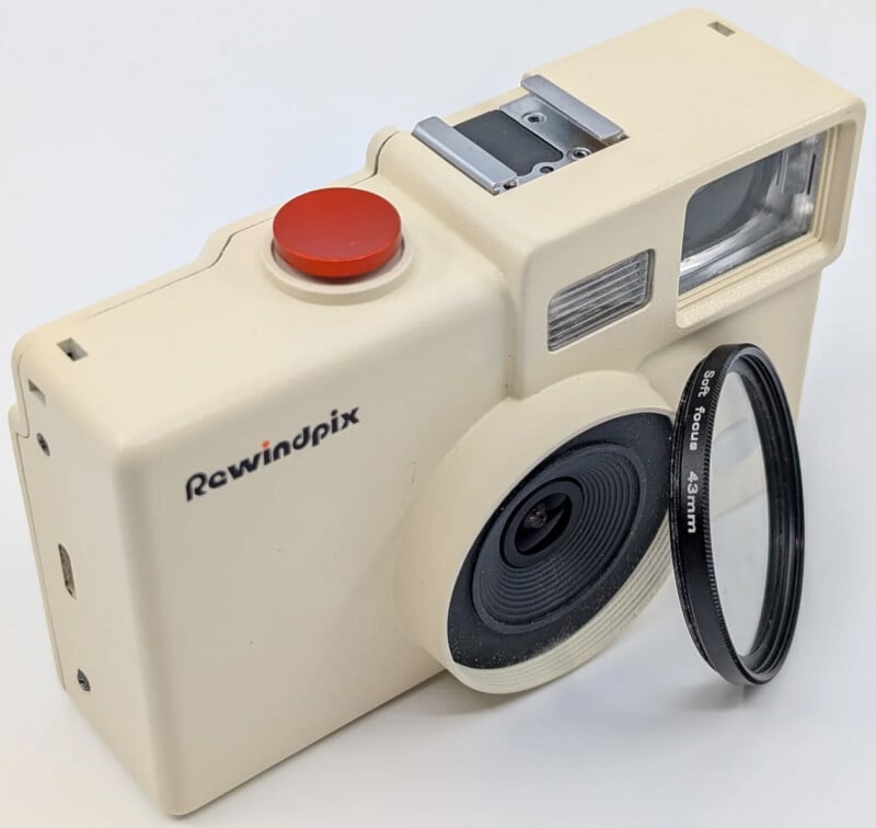 Una cámara Rewindpix de color crema mostrada sobre una superficie blanca, con un gran botón obturador rojo, una lente sobresaliente y un filtro extraíble de 43 mm montado en el frente.