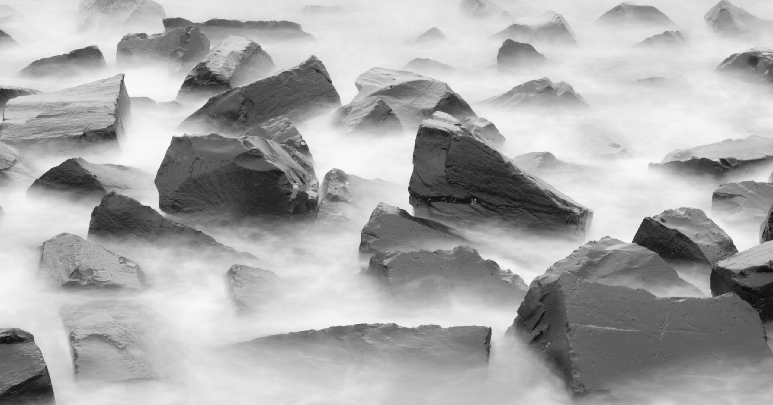 Enormes rocas irregulares están parcialmente sumergidas en agua brumosa que fluye, creando una escena en blanco y negro surrealista y etérea.