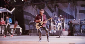 A guitarist in a maroon jacket plays an electric guitar on stage with a band; other musicians and instruments are visible in the background, with lights and equipment around them.
