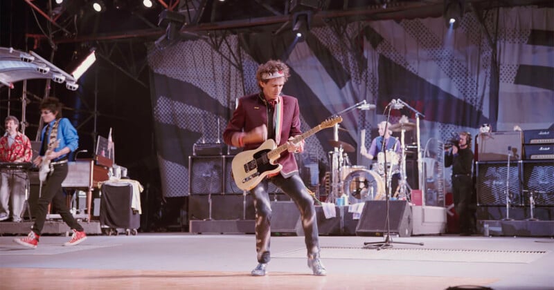 A guitarist in a maroon jacket plays an electric guitar on stage with a band; other musicians and instruments are visible in the background, with lights and equipment around them.