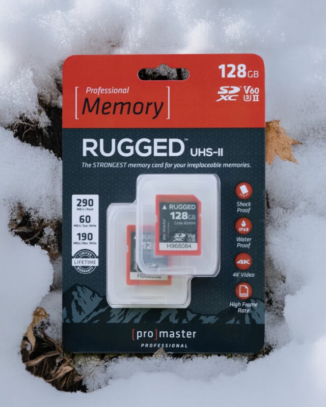 A ProMaster Professional 128GB Rugged UHS-II SD card in packaging lies on snow. The package highlights features like shockproof, waterproof, 4K video support, and high write speed. The SD card is visible inside its case.