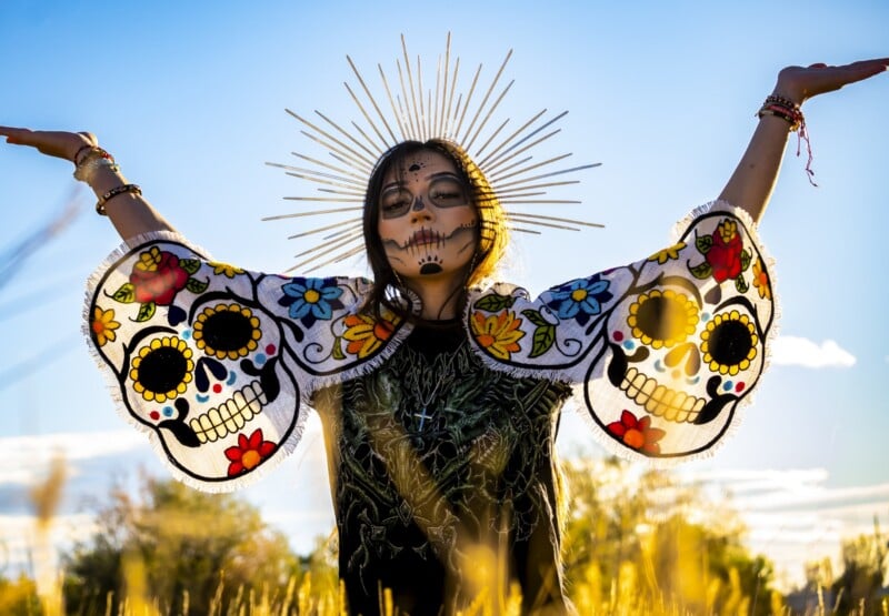 A person with face painted like a sugar skull, wearing a black outfit with colorful skull designs on wide sleeves, stands in a field with arms raised. Gold rays form a halo around their head, evoking Día de los Muertos.