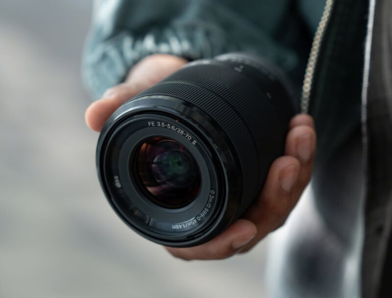 Una persona sostiene la lente de una cámara en su mano derecha, con la lente mirando al espectador. El texto en la lente dice "FE 3,5-5,6/28-70." El fondo está borroso.