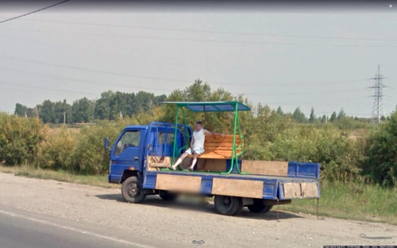 Un camión de plataforma azul estaba estacionado al costado de la carretera, con un banco de madera y un refugio sobre la plataforma. Hay un hombre sentado en el banco. Al fondo se ven árboles y líneas eléctricas.