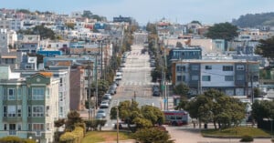 A wide, steep street runs through a residential neighborhood with colorful houses, trees, and power lines on both sides. A bus and several cars are visible on the street, with a park area in the foreground.