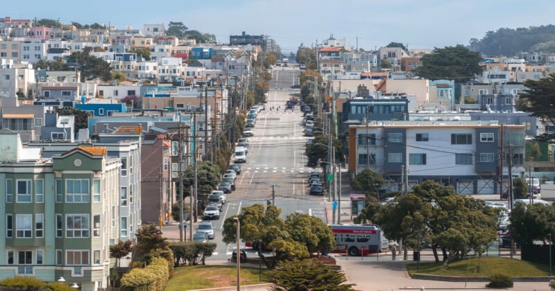 Una calle ancha y empinada atraviesa la zona residencial, bordeada de casas coloridas, árboles y líneas eléctricas. En la calle se ven un autobús y varios coches, y en primer plano una zona de parque.