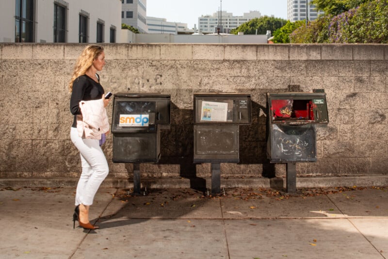 A woman in white pants and black heels walks past three weathered newspaper dispensers on a city sidewalk, next to a concrete wall, holding a white jacket and cellphone.