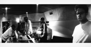 Black and white photo of several people in a recording studio. Some are standing by a keyboard and laptops, one person sits on a couch, and another stands in the foreground, looking towards the camera.