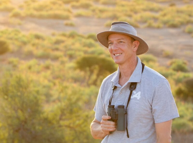 Un hombre sonriente con un sombrero gris y una camiseta polo está parado al aire libre con binoculares frente a un soleado paisaje de hierba al fondo.