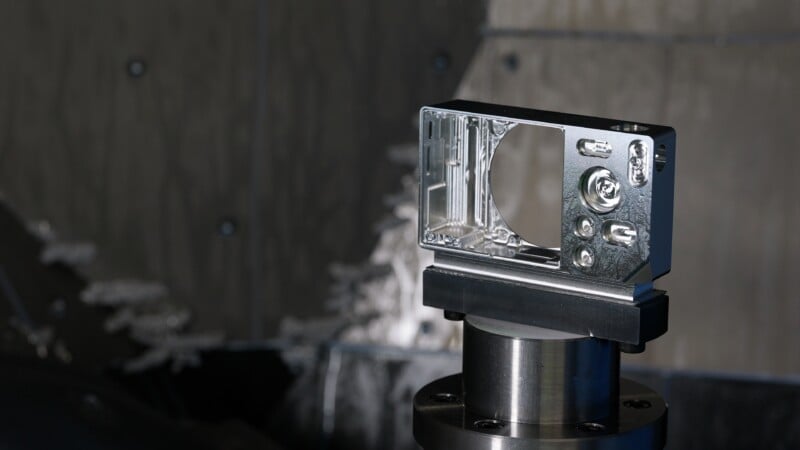 A machined metal component with precise cutouts is mounted on a cylindrical fixture, set against a blurred industrial background with gray tones.