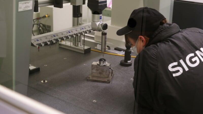 A person wearing a jacket labeled “SIGN” is closely inspecting a metal component on a precision measuring machine inside a laboratory or industrial setting.