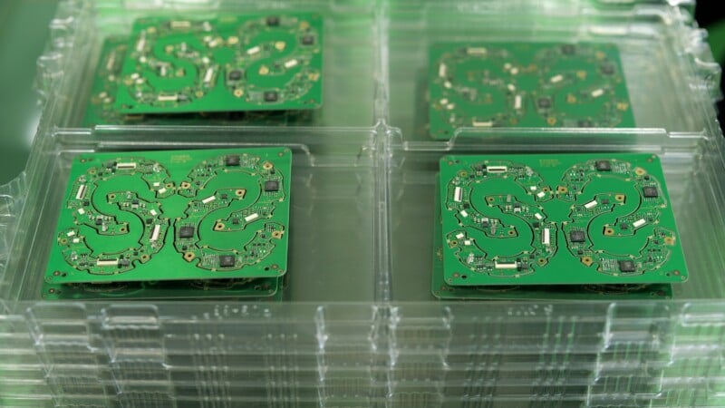Four green square circuit boards with intricate pathways are stacked in clear plastic trays, arranged in a 2x2 layout, likely prepared for assembly or inspection in an electronics manufacturing setting.