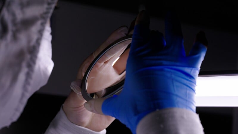 A person wearing a white lab coat and blue gloves inspects a circular glass object under bright light, suggesting a scientific or cleanroom environment.