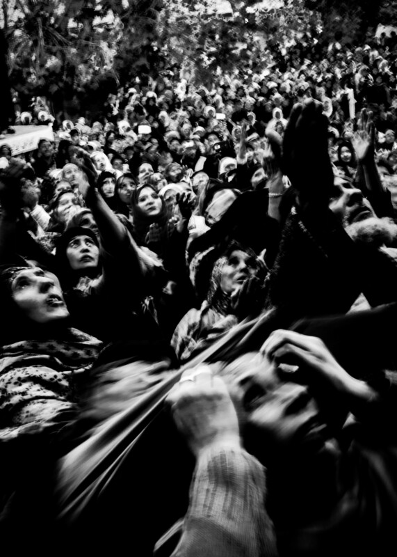 A large crowd of people, mostly women in headscarves, reach upwards with anxious expressions. The black and white image is blurred, conveying a sense of urgency and emotion in the densely packed group.