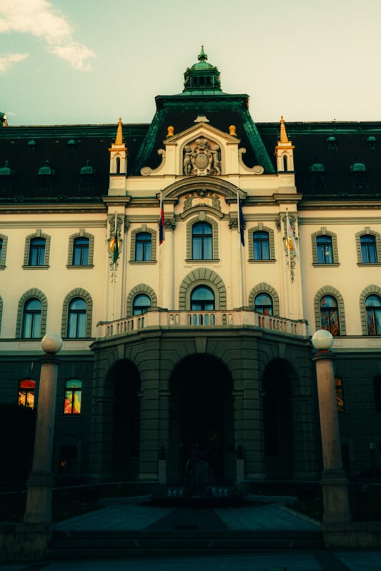 Un gran edificio histórico con una entrada arqueada, decoración ornamentada y techo de tejas verdes, fotografiado al atardecer con un cielo cambiante de fondo. La entrada en primer plano está enmarcada por columnas.
