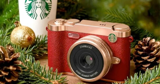 A red and gold camera with a Starbucks logo sits among pine branches, pinecones, and a gold ornament. A Starbucks cup is in the background, creating a festive holiday setting.