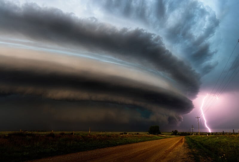 Una enorme y oscura nube de supercélulas dominaba el cielo sobre un camino de tierra rural, mientras un brillante rayo caía cerca de líneas eléctricas en la distancia.