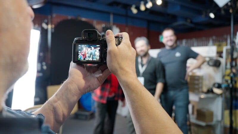 Primer plano de una persona sosteniendo una cámara, capturando a tres personas posando y sonriendo en un ambiente interior informal con luces de estudio y equipo de fondo.