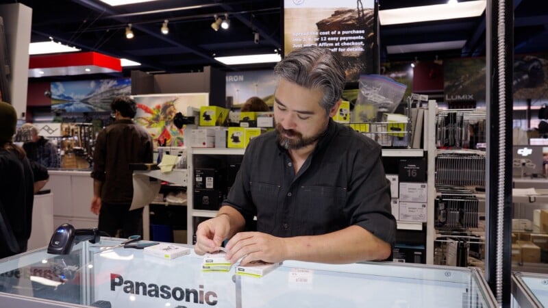Un hombre barbudo y de pelo gris se encuentra detrás del mostrador de Panasonic en una tienda de electrónica, sosteniendo una pequeña caja de producto. Al fondo se pueden ver estantes que contienen equipos y accesorios para cámaras.