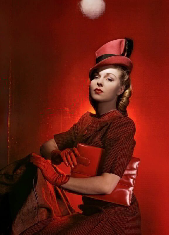 Una mujer vestida con un traje rojo vintage, sombrero, guantes y bolso de mano posa sobre un fondo rojo brillante, exudando elegancia y aplomo.