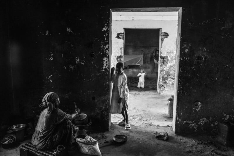 Una fotografía en blanco y negro muestra a una mujer sentada en el suelo cerca de una pared preparando comida, un niño de pie en la puerta y otro más atrás en una habitación separada y escasamente amueblada.