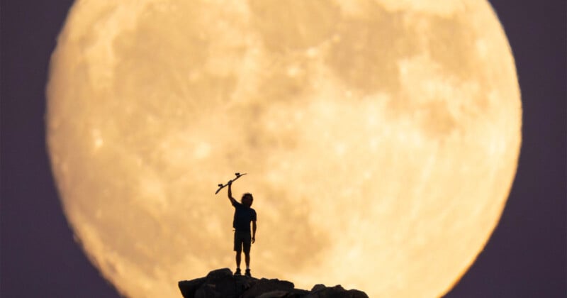 A person stands on a rocky hilltop holding an object above their head, silhouetted against a giant, bright full moon in the background.