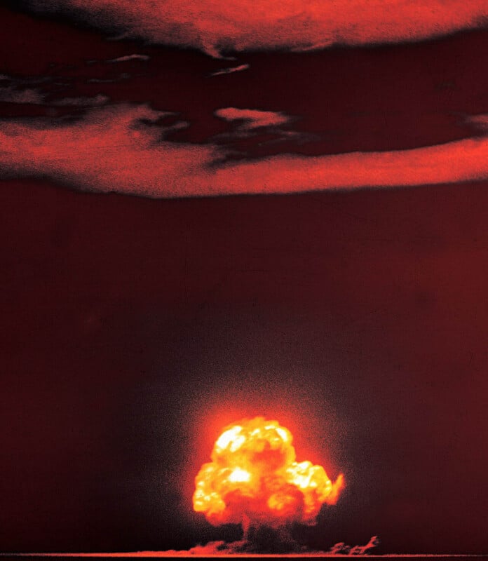 A large, bright explosion creates a mushroom cloud against a dark red sky, with scattered clouds above and a glowing horizon below.