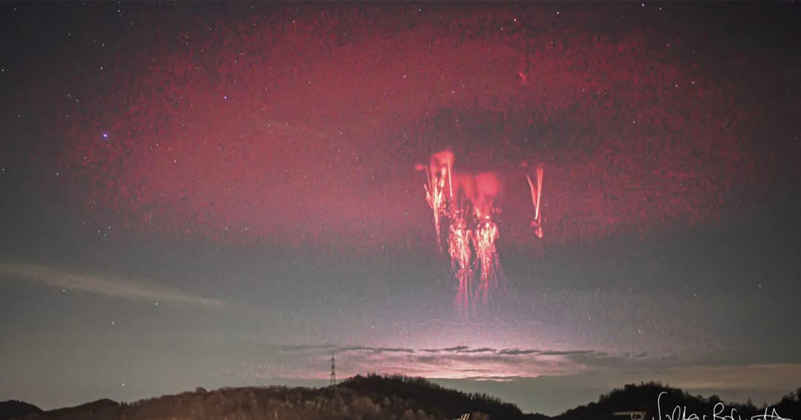 A bright red jellyfish-shaped lightning, known as a sprite, illuminates the night sky above a dark landscape with clouds and faint stars visible in the background.