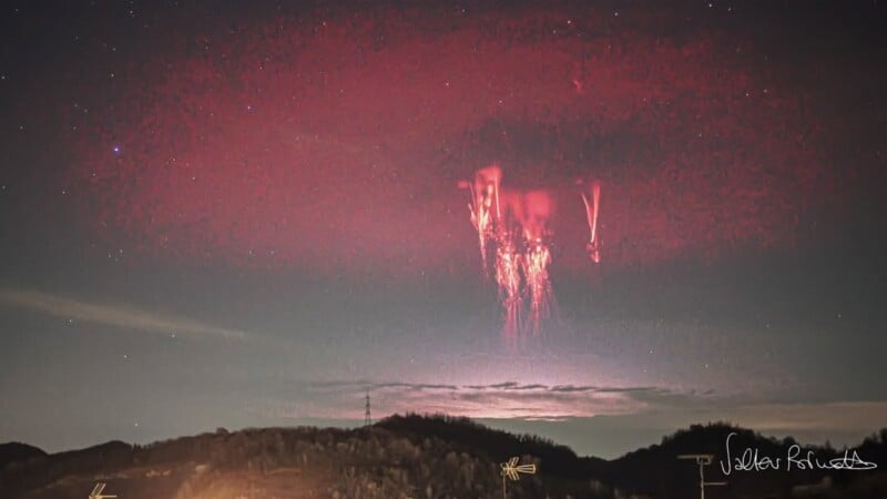El cielo nocturno presenta un raro fenómeno de relámpagos de elfos rojos en el horizonte distante. Formas brillantes de color rojo y naranja aparecen en lo alto de las colinas oscuras, contrastando con un cielo de estrellas tenues.