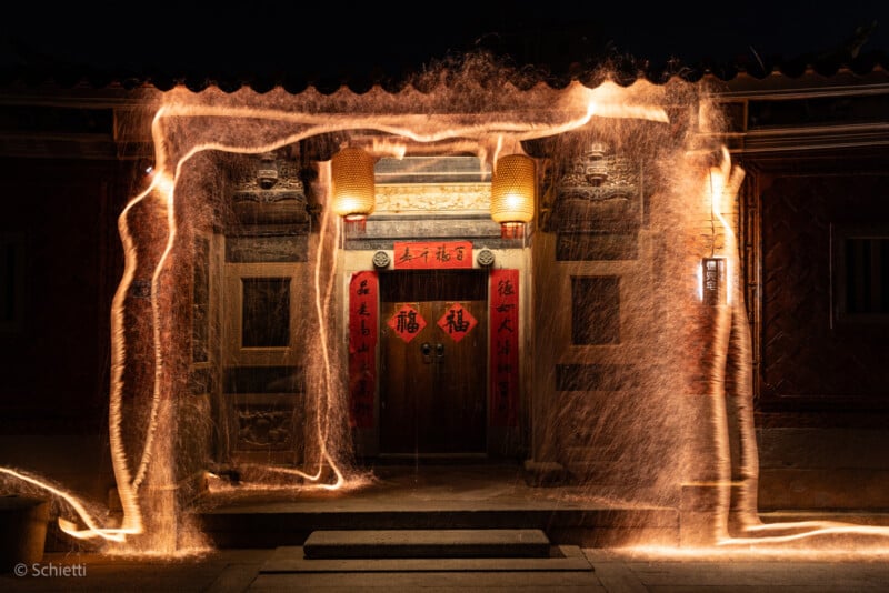 Entrada tradicional china con faroles rojos y pancartas de caligrafía, muy iluminada por la noche. Senderos de luz dorada se arremolinaban, creando patrones brillantes frente a la puerta.