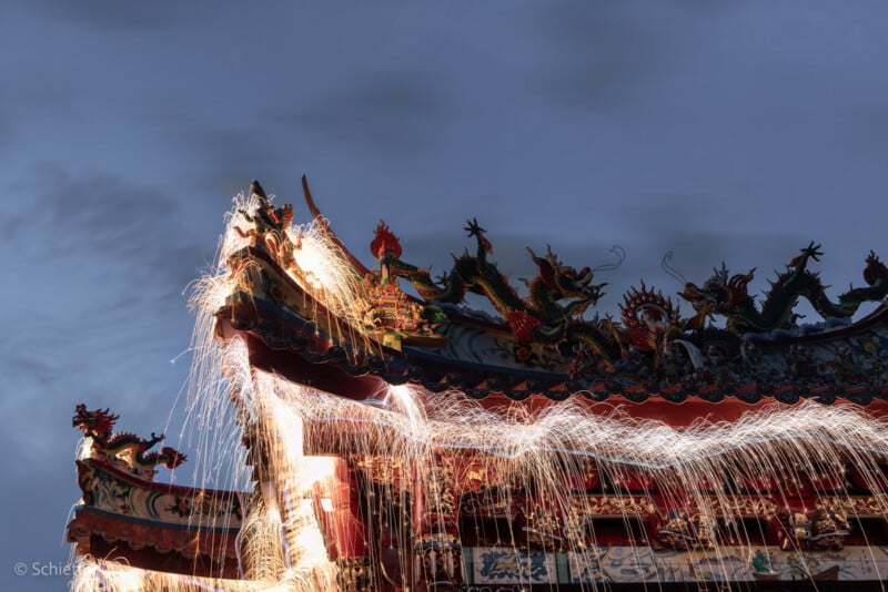 Contra el cielo oscuro, los fuegos artificiales lanzaban chispas desde el techo de un templo tradicional asiático decorado con ornamentadas esculturas de dragones. Las exposiciones prolongadas crean estelas de luz que recorren el techo.