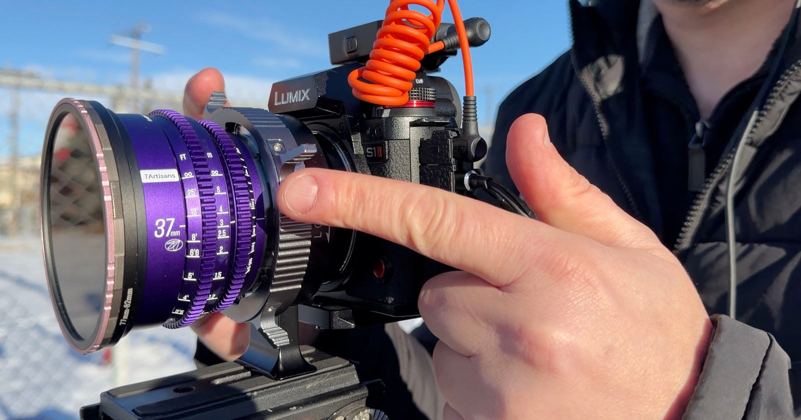 A person adjusts the purple focus ring on a LUMIX camera with an external microphone attached, outdoors on a sunny day.