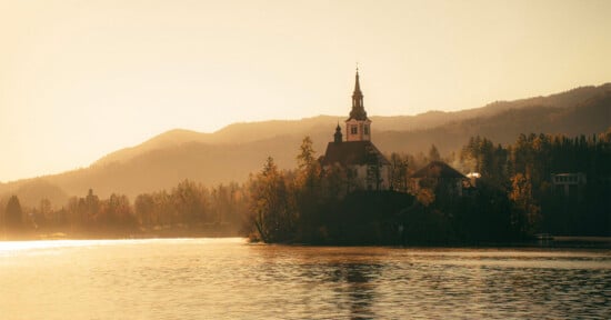 A church with a tall steeple stands on an island surrounded by a calm lake, with forested hills and mountains in the background at sunset. The scene is bathed in warm golden light.