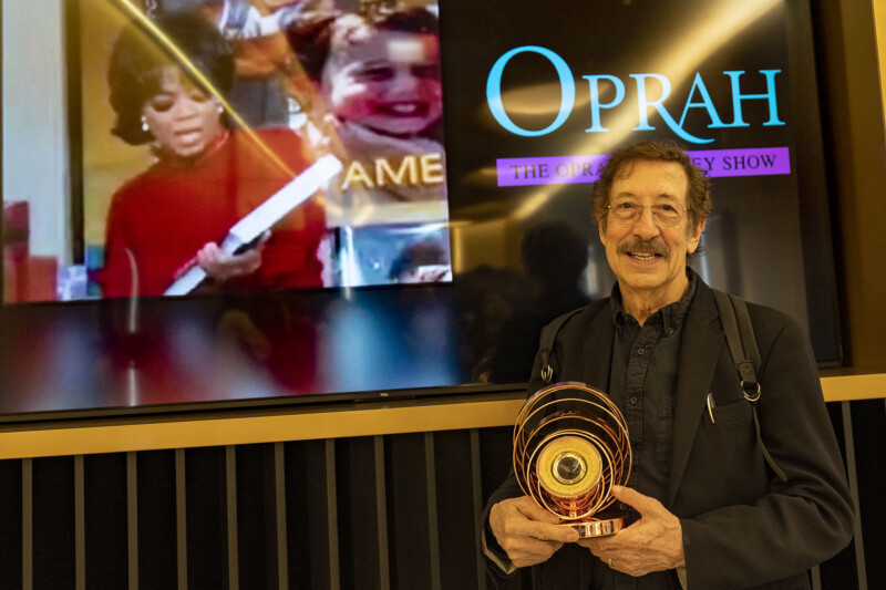 A man with gray hair and glasses smiles while holding a circular award. Behind him, a screen displays “The Oprah Winfrey Show” logo and an image of Oprah Winfrey holding a large object.