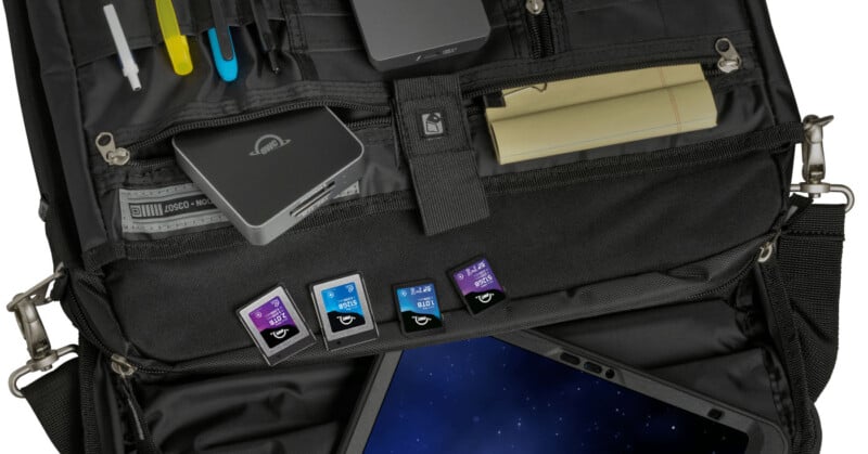 Al abrir la bolsa tecnológica negra se revela un bolígrafo, un bloc de notas, cables, varias tarjetas SD en las ranuras y una tableta parcialmente visible en un compartimento acolchado. En la bolsa se encuentran cuidadosamente ordenados varios accesorios electrónicos.