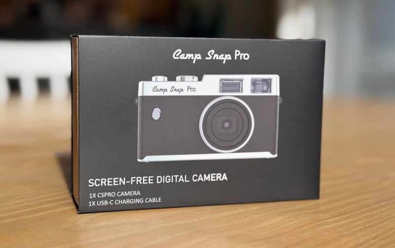 A black box of the Camp Snap Pro screen-free digital camera sits on a wooden surface. The box shows an image of the camera and lists contents: 1x CSPRO camera and 1x USB-C charging cable.
