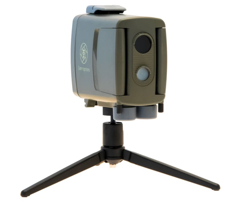 A compact, green and gray trail camera with a lens and motion sensor on the front, mounted on a small black tripod with three legs splayed outward.