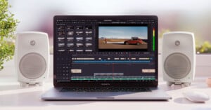 A MacBook Pro on a desk displays video editing software with a car video. Two white speakers are on either side of the laptop, and sunlight streams in through a window. A small plant is visible on the left.