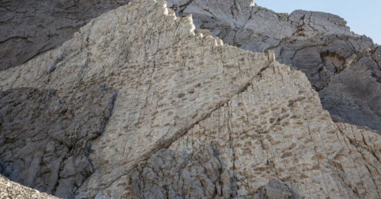 Large rocky cliff face with visible diagonal cracks and numerous small, round indentations covering some of the lighter rock surface; the background shows more rugged gray stone.