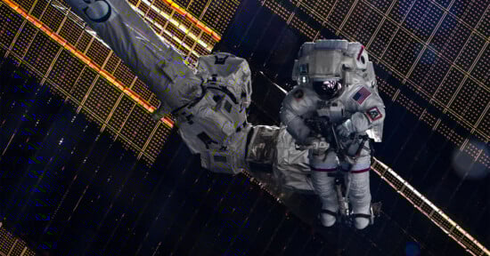 An astronaut in a spacesuit works outside the International Space Station, surrounded by large solar panels and equipment, with the darkness of space in the background.