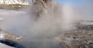 A geyser erupts, shooting steam and water into the air amidst a rocky landscape with snow-covered trees and hills in the background.