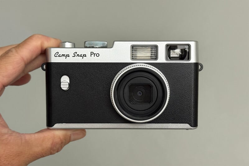 A hand holds a black and silver vintage-style camera labeled "Camp Snap Pro" against a plain gray background.