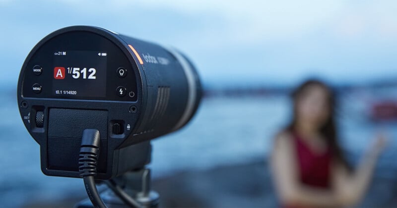 Primer plano del flash de una cámara profesional que muestra la configuración digital, ubicado cerca del agua al aire libre. En el fondo borroso, un hombre vestido de rojo se encuentra cerca de la costa.