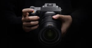 A person in a black outfit holds a large Hasselblad camera with both hands, focusing on the camera’s lens and controls against a dark background.
