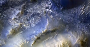A high-resolution satellite image shows a rugged, rocky landscape with ridges, small dunes, and varying shades of blue, gray, and tan, resembling a Martian surface with textured terrain and subtle elevation changes.