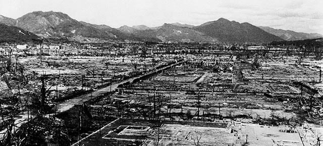 A black-and-white photo showing a vast, devastated landscape with ruins and debris. Bare mountains rise in the background, and little remains of buildings, illustrating the aftermath of destruction.