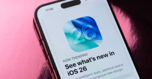 A smartphone screen displays an app update notification for iOS 26, featuring a blue and green abstract icon with the number 26 and the headline “See what’s new in iOS 26.” The background is pink and shiny.