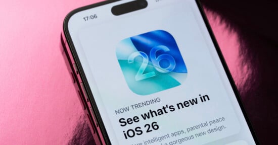 A smartphone screen displays an app update notification for iOS 26, featuring a blue and green abstract icon with the number 26 and the headline “See what’s new in iOS 26.” The background is pink and shiny.